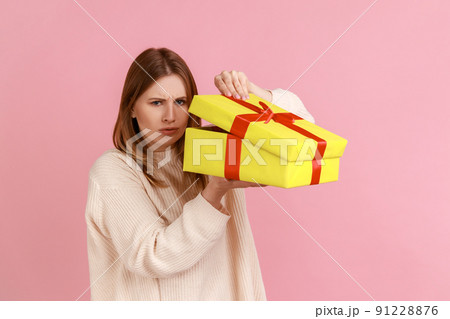 Portrait of disappointed young adult blond woman opening wrapped present box, does not like her gift, fells sorrow, wearing white sweater. Indoor studio shot isolated on pink background. 91228876