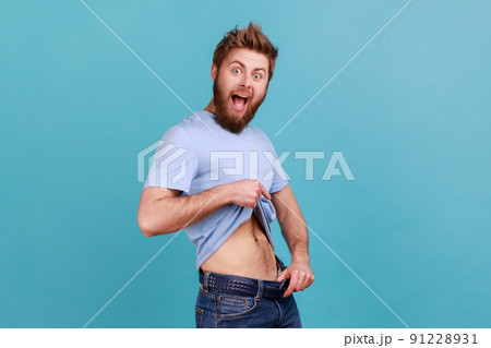 Portrait of bearded man having willpower points at slim waist in big trousers, successful weight loss, keeping diet and going for sport. Indoor studio shot isolated on blue background. Portrait of bearded man having willpower points at slim waist in big trousers, successful weight loss, keeping diet and going for sport. Indoor studio shot isolated on blue background. 91228931