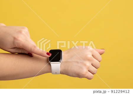 Closeup side view of woman hand with smartwatch touching touchscreen, arm with wristtwatch with white strap, technology device. Indoor studio shot isolated on yellow background. 91229412