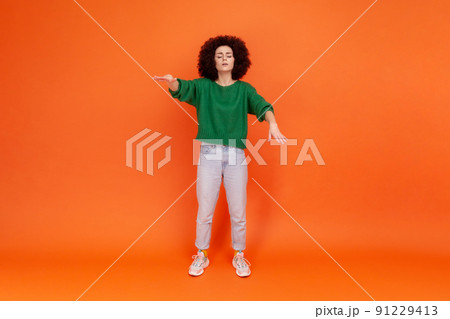 Full length portrait of woman with Afro hairstyle in green sweater walking with closed eyes, stretching hands to search way, vision problems. Indoor studio shot isolated on orange background. 91229413