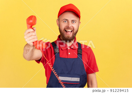 Portrait of positive handyman wearing blue uniform holding out handset, asking call him and order service, looking at camera happy expression. Indoor studio shot isolated on yellow background. 91229415