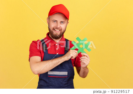 Portrait of positive delighted workman wearing blue overalls standing, holding green hashtag, looking at camera with happy expression. Indoor studio shot isolated on yellow background. 91229517