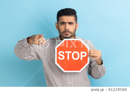Portrait of serious business holding big red stop road sign and pointing finger on you, strictly looking at camera, wearing striped shirt. Indoor studio shot isolated on blue background. 91229560
