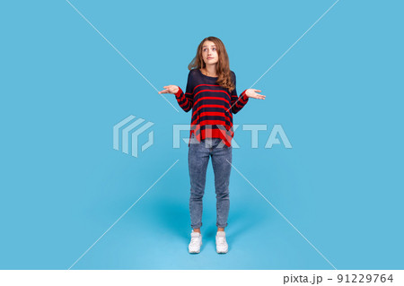 Full length portrait of uncertain woman wearing striped sweater looking desperate, making helpless gesture with her hands, doesn't know what to do. Indoor studio shot isolated on blue background. 91229764