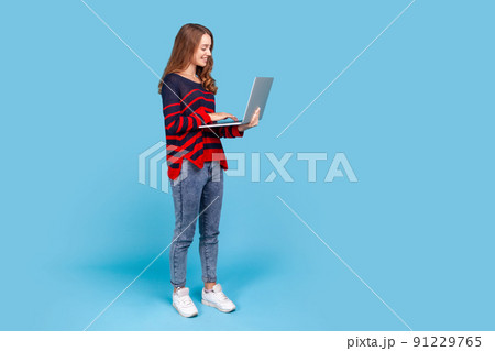 Full length portrait of charming woman in sweater, standing with portable computer in hands, looking at laptop screen with smile, satisfied with work. Indoor studio shot isolated on blue background. 91229765
