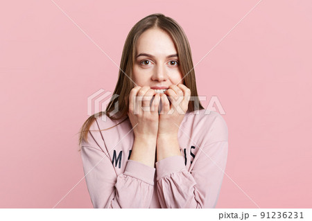 Isolated shot of pretty female model bites nails as feels very nervous and embarrassed, worries before important event, isolated over pink background. Frightened young woman looks anxiously at camera 91236231