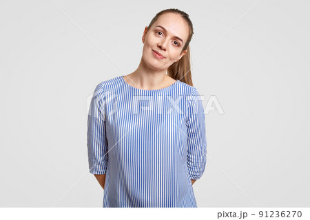 Beautiful teenage girl has dark hair combed in pony tail, wears striped blue shirt, keeps hands behind back, looks directly at camera, isolated over white background. People and beauty concept 91236270