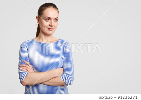 Horizontal shot of thoughtful European female with pony tail, keeps hands crossed, dressed in striped blouse, poses against white background with copy space aside for your promotion or advertisement 91236271