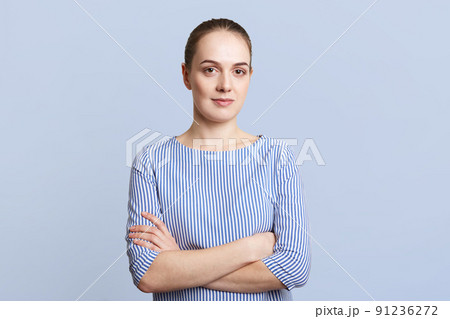 Pretty brunette female wears striped elegant blouse, keeps arms folded, has serious expression, listens to interlocutor attentively, isolated over blue background. People, facial expressions concept 91236272