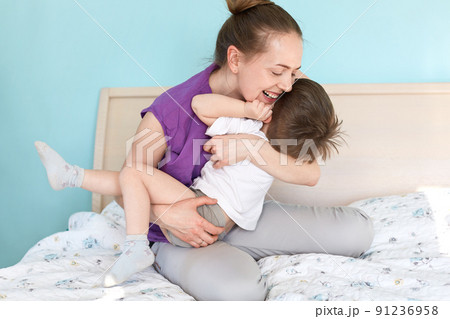 Positive young attractive mother play with her little son, teases and expresses happiness, have joy after good sleep, sit on soft bed, enjoys calm domestic atmosphere at home. Motherhood concept Positive young attractive mother play with her little son, teases and expresses happiness, have joy after good sleep, sit on soft bed, enjoys calm domestic atmosphere at home. Motherhood concept 91236958
