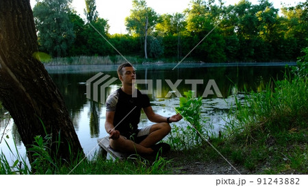 Sporty handsome caucasian young man working out on lake in park, sitting in squat, doing Half Lotus, Padangustha Padma Utkatasana, hands in Namaste, full length 91243882