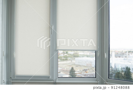 Details of white fabric roller blinds on the plastic window with wood texture in the living room. Details of white fabric roller blinds on the plastic window with wood texture in the living room. 91248088