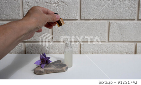 A bottle of serum and a hand with a pipette on a white brick background. A close-up of a frosted glass container for a cosmetic skin care product. A bottle of serum and a hand with a pipette on a white brick background. A close-up of a frosted glass container for a cosmetic skin care product. 91251742