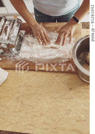 A woman's hands roll out a piece of noodle dough on a wooden board covered with flour. 91261042