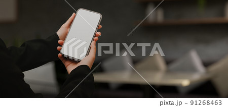 A female hands holding a mobile phone white screen mockup over blurred modern living room 91268463