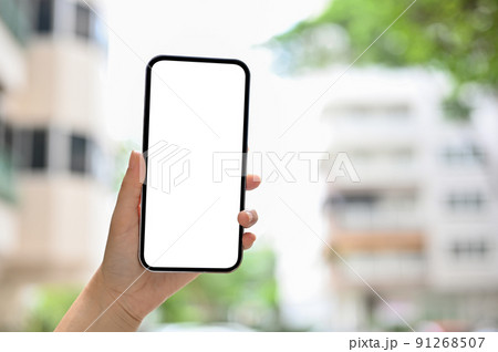 A smartphone white screen mockup in a female's hand over blurred flat or apartment building 91268507