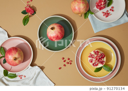 Shot of colorful plates with Fresh pomegranates, ripe seeds whole pomegranates, opened pomegranate, green leaves  and linen napkins on brown table. Top view for design. Dessert fruit 91270134