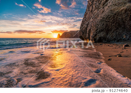 Bright sea sunset. The waves crash into the rock, lit by the warm sunset, sand and pebbles, volcanic basalt as in Iceland. Sea wave breaks into splashes and white foam. Never-ending beauty of nature. 91274640