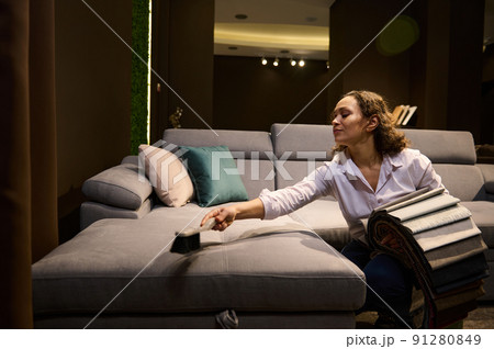 Beautiful Hispanic woman- inspired interior designer, professional decorator holding upholstery fabric swatches and cleaning the velour surface of a gray art nouveau sofa in the home interior Beautiful Hispanic woman- inspired interior designer, professional decorator holding upholstery fabric swatches and cleaning the velour surface of a gray art nouveau sofa in the home interior 91280849