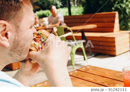 Young hungry caucasian man eating hamburger or beef burger with vegetables and sauce in outdoor street cafe. Having brunch on sunny day. Close-up, fastfood, portrait. Young hungry caucasian man eating hamburger or beef burger with vegetables and sauce in outdoor street cafe. Having brunch on sunny day. Close-up, fastfood, portrait. 91282131