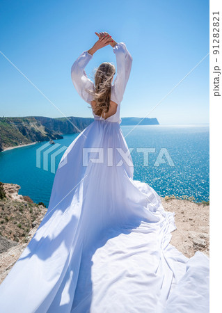 Blonde with long hair on a sunny seashore in a white flowing dress, rear view, silk fabric waving in the wind. Against the backdrop of the blue sky and mountains on the seashore. Blonde with long hair on a sunny seashore in a white flowing dress, rear view, silk fabric waving in the wind. Against the backdrop of the blue sky and mountains on the seashore. 91282821
