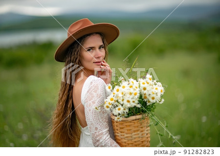 A middle-aged woman in a white dress and brown hat stands on a green field and holds a basket in her hands with a large bouquet of daisies. In the background there are mountains and a lake. A middle-aged woman in a white dress and brown hat stands on a green field and holds a basket in her hands with a large bouquet of daisies. In the background there are mountains and a lake. 91282823