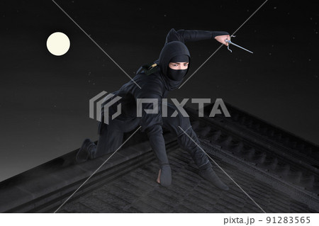 満月の夜の暗闇に家屋の屋根の上で一人の黒い服装の忍者が武器を持って移動する 91283565