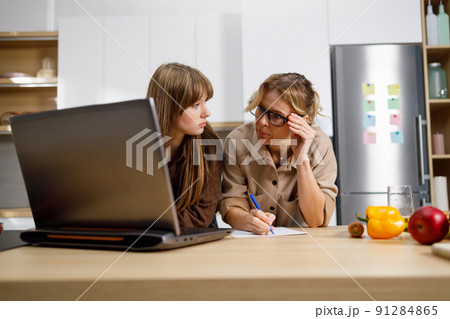 Adult woman and her daughter in the kitchen writing something while sitting in front of a laptop 91284865
