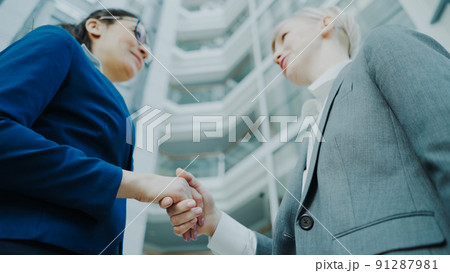 Low angle shot of businesswoman shaking hands and talking with female business colleague in suit in hall of modern office building 91287981