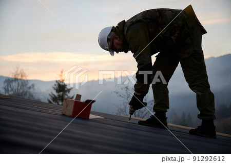 Male builder installing black corrugated iron sheet used as facade of future cottage. Silhouette of man worker building wooden frame house. Carpentry and construction concept. 91292612