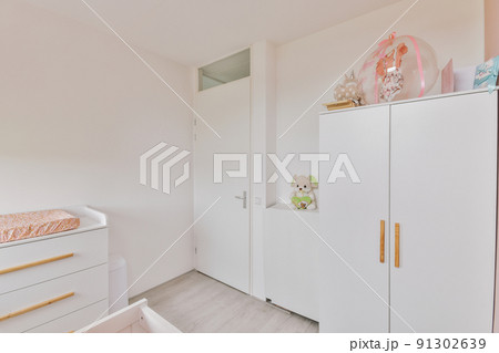 Interior of white room with baby crib and armchair near window with blue chest of drawers in daylight Interior of white room with baby crib and armchair near window with blue chest of drawers in daylight 91302639
