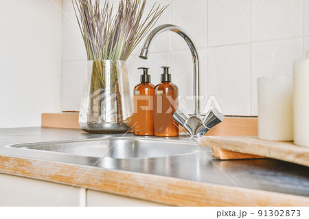 A fragment of the interior of a light home kitchen with beige furniture in a minimalist style with a sink, mixer tap, soap and candles 91302873