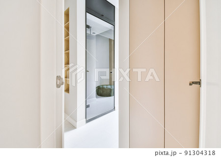 Narrow corridor with white walls and a door leading to a spacious room with a mirror and parquet flooring in a modern apartment 91304318