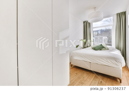 Comfortable bed and wardrobe placed in small narrow minimalist style bedroom with white walls and green curtains on window in modern apartment Comfortable bed and wardrobe placed in small narrow minimalist style bedroom with white walls and green curtains on window in modern apartment 91306237