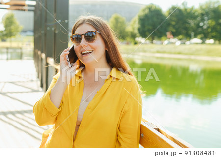 Smiling beautiful woman talking on phone on river background. Happy woman with yellow shopping bag is using a smartphone outdoors, summer time. Traveler. Women posed against nature park Smiling beautiful woman talking on phone on river background. Happy woman with yellow shopping bag is using a smartphone outdoors, summer time. Traveler. Women posed against nature park 91308481