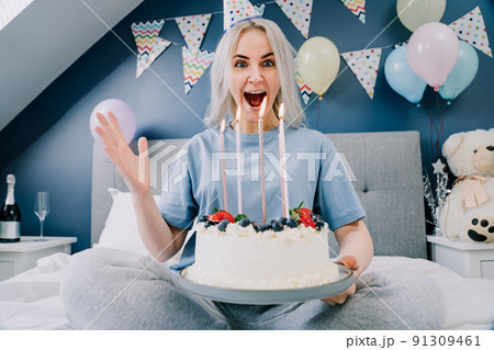 Excited emotional woman in birthday cap holding festive cake with candles, while sitting on the bed in decorated bedroom. Morning surprise. Happy birthday concept. Selective focus. Excited emotional woman in birthday cap holding festive cake with candles, while sitting on the bed in decorated bedroom. Morning surprise. Happy birthday concept. Selective focus. 91309461
