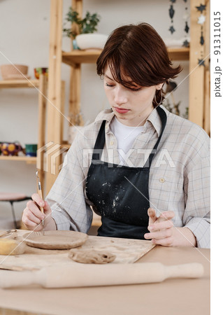Brunette female in apron sitting by workplace and using wooden cutting handtool Brunette female in apron sitting by workplace and using wooden cutting handtool 91311015
