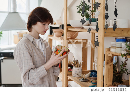 Young brunette woman choosing gift in earthenware shop while holding clay bowl 91311052