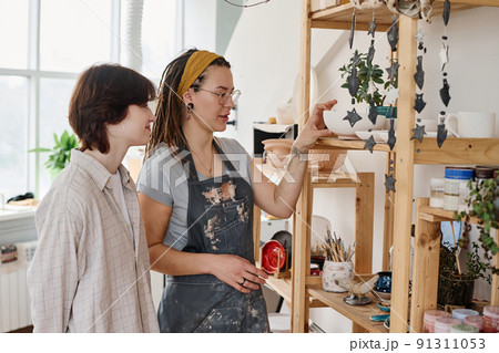 Young female clerk of small earthenware shop showing new items to customer 91311053