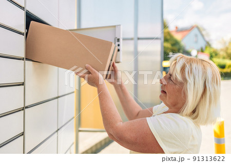 50s caucasian woman getting parcel from cell of automatic post terminal outdoors. 50s caucasian woman getting parcel from cell of automatic post terminal outdoors. 91313622