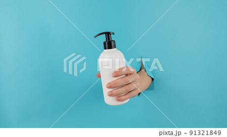 A woman's hand sticks out of a hole in a blue paper background and holds a skin lotion pump. A woman's hand sticks out of a hole in a blue paper background and holds a skin lotion pump. 91321849