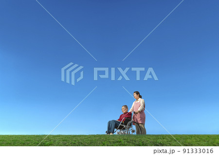 青空を背景に車椅子に乗った高齢の女性を介護する中年女性。家族、高齢、介護のイメージ 青空を背景に車椅子に乗った高齢の女性を介護する中年女性。家族、高齢、介護のイメージ 91333016