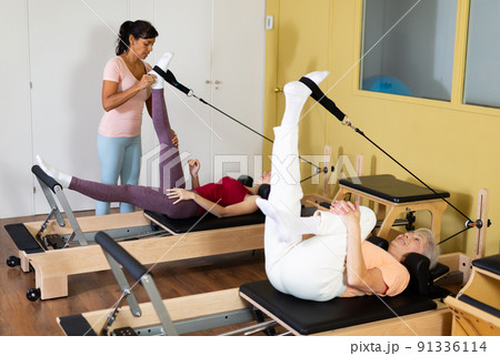 Active women perform Pilates exercises on a simulator with the help of a trainer 91336114
