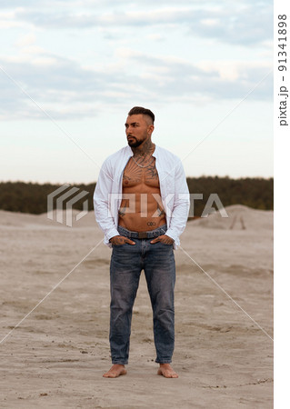 Muscular athletic sexy man in white shirt with bare-chested rests on beach. Portrait of tanned fit male model on vacation 91341898