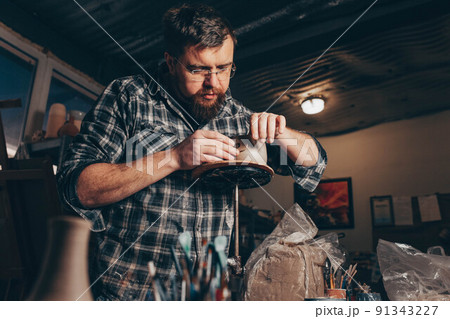 Bearded man works in a pottery workshop, concentrating on the process of creating a clay cup. Bearded man works in a pottery workshop, concentrating on the process of creating a clay cup. 91343227