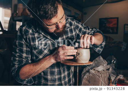 Bearded man works in a pottery workshop, concentrating on the process of creating a clay cup. 91343228