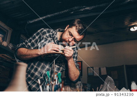 Bearded man works in a pottery workshop, concentrating on the process of creating a clay cup. 91343230