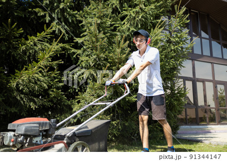 Young man mows a lawn near a large mansion. Worker with a gasoline lawn mower 91344147