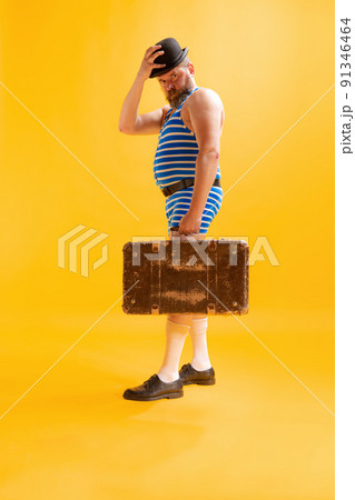 One funny beachgoer, fat excited man wearing retro striped swimsuit and vintage bowler hat holding old suitcase isolated on bright yellow background. Vacation, summer, humor One funny beachgoer, fat excited man wearing retro striped swimsuit and vintage bowler hat holding old suitcase isolated on bright yellow background. Vacation, summer, humor 91346464