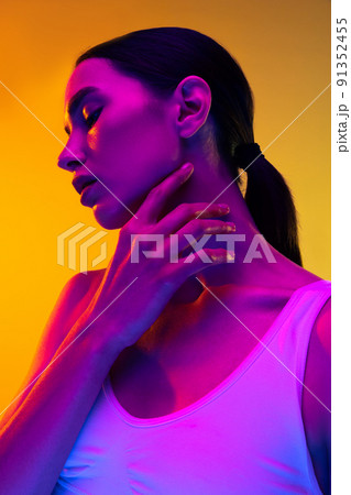 Portrait of tender young girl wit nude makeup and low pony rail posing isolated over yellow studio background in neon light 91352455